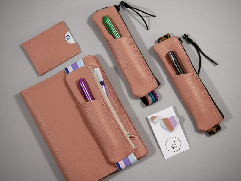 carnet cuir A5 présenté avec différents modèles de trousses élastiques coordonnées permettant de le maintenir fermé, et porte-cartes et pin's dans les même tons