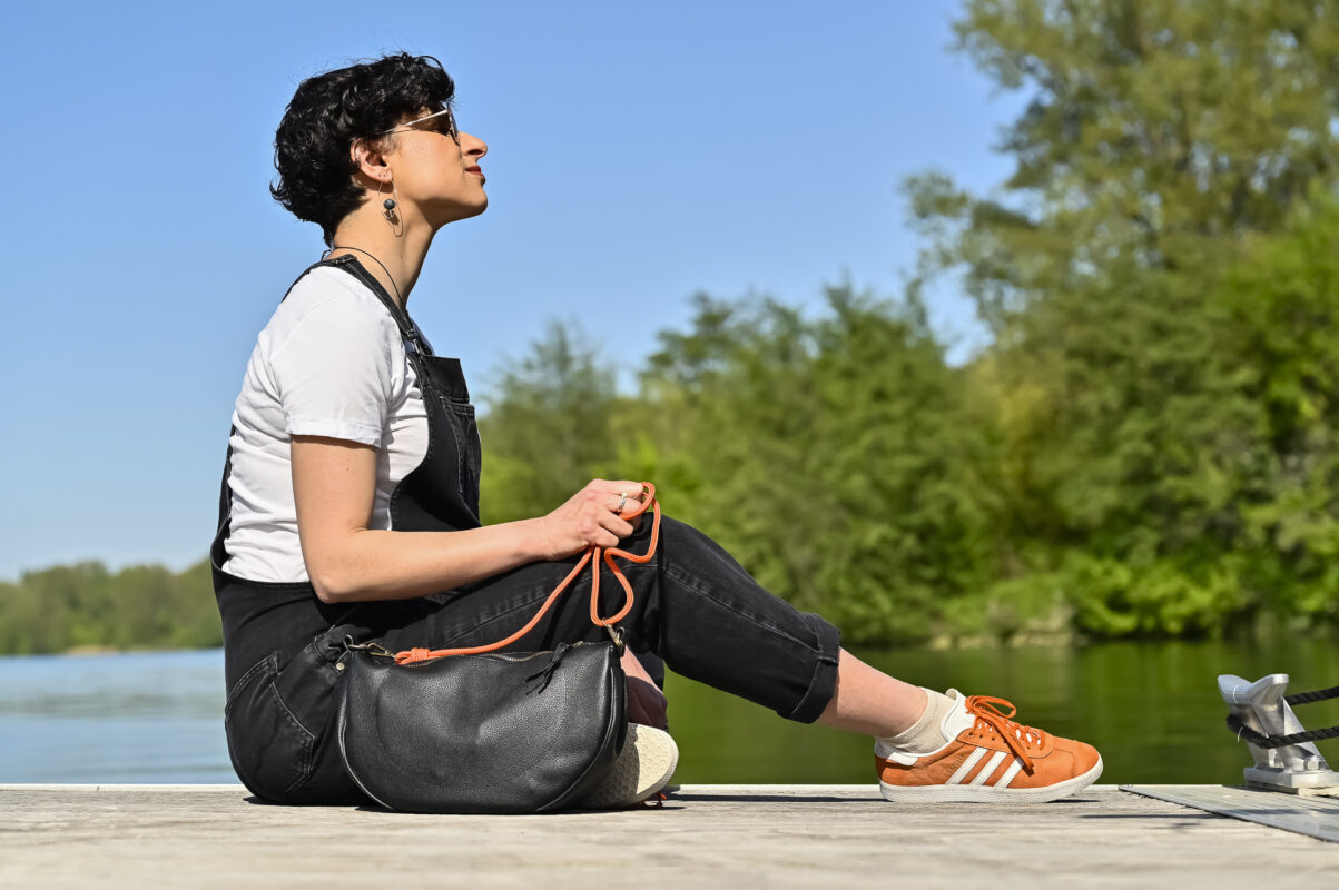 Sac demi-lune en cuir noir avec sangle en corde contrasté orange, posé sur un pont de bateau à côté du mannequin assis