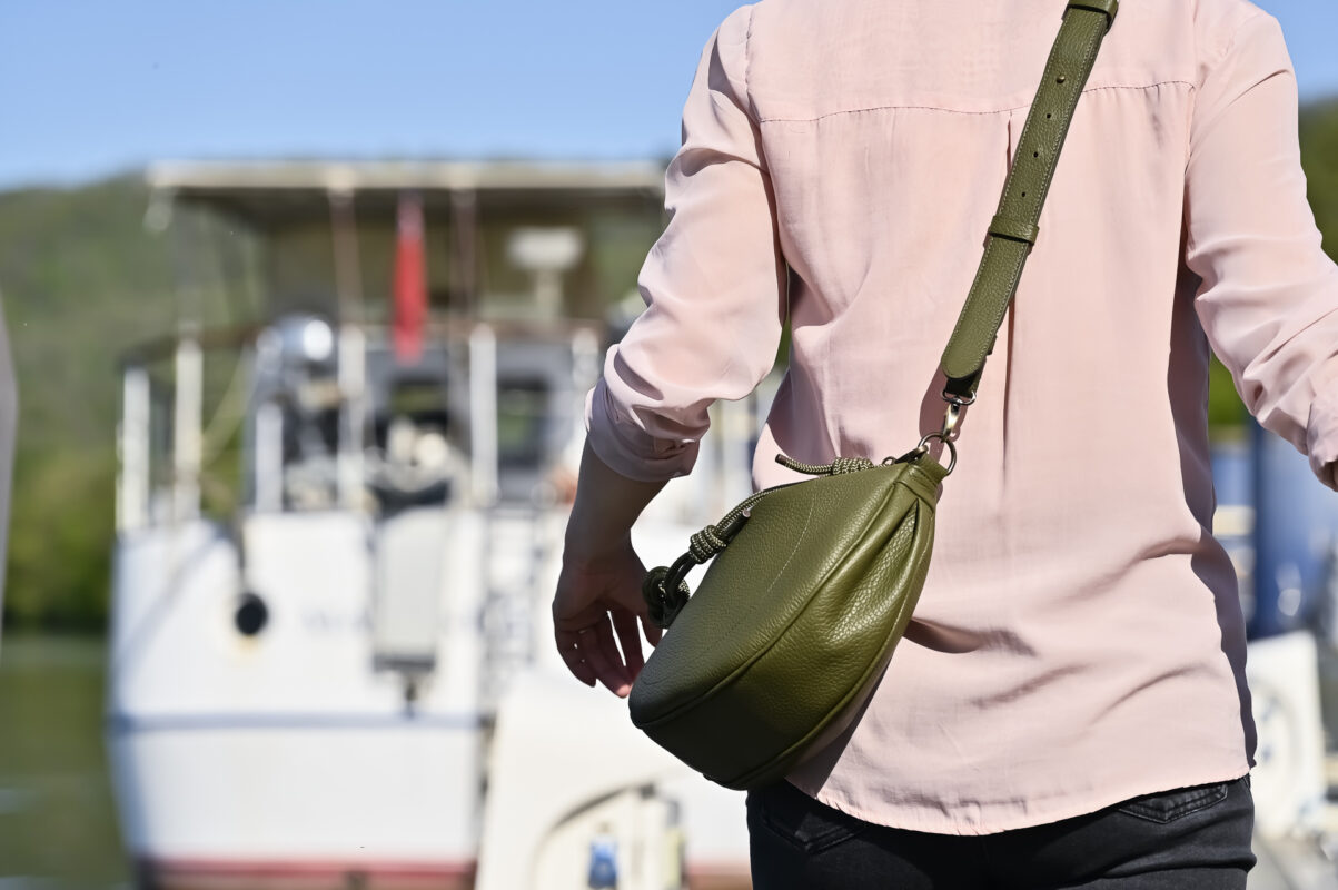 Sac demi-lune en cuir kaki avec sangles en cuir, porté en bandoulière par le mannequin, dans un port de plaisance