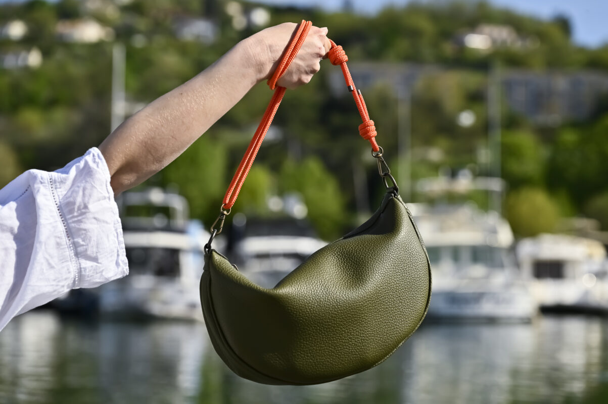 Sac demi-lune en cuir kaki avec sangles en corde orange contrastée, porté à la main par le mannequin, dans un port de plaisance